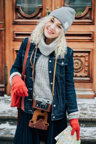 Joven turista rubia rizada en ropa de abrigo con cámara de película vieja y mapa de la ciudad de pie sobre el fondo de puertas de madera, invierno, nieve —  Fotos de Stock