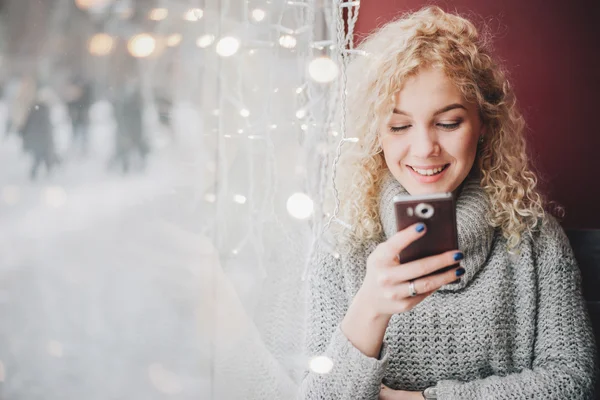 Молодая блондинка кудрявая в теплом свитере с помощью смартфона и улыбаясь в кафе, зимний город за окном Стоковое Фото