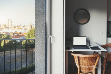 Masasında dizüstü bilgisayarı, grafik tableti, kalemi, akıllı telefonu ve USB merkezi olan rahat bir dairede bir masaüstü, iki baston sandalye, pencerenin dışında manzaralı bir balkon.