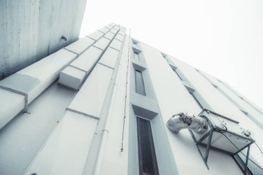 Solunda üç dikey havalandırma kanalı, tüp ve bir paratoner bulunan beyaz bir binanın ön cephesinin geniş açılı alt görüntüsü, bir grup uzamış pencere ve harici bir klima modülü.