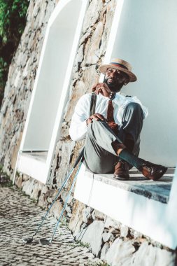 Eski bir kostüm ve şapka giymiş düşünceli bir siyah retroseksüel adam elinde bir puro ve yanında iki golf sopasıyla bir binanın teneffüsünde dinleniyor ve oturuyor.