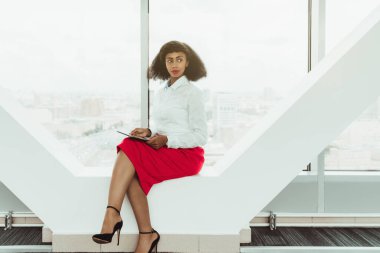 Büyüleyici, narin, Afro-Amerikalı iş kadını modern ofisin çağdaş bir vitrininde elinde dijital tabletle oturmakta.