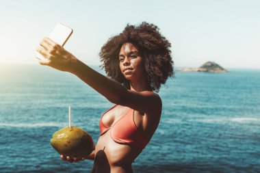 Elinde samanla ve okyanusla bir yat güvertesinde dururken akıllı telefonuyla selfie çeken muhteşem bir Afro-Amerikan kadının portresi.