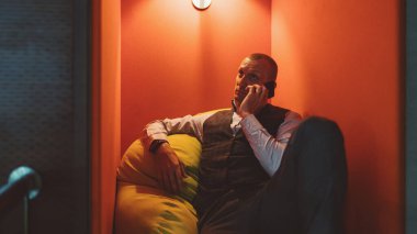 Yakışıklı, olgun, beyaz bir girişimci, kafasında fenerle modern bir ofisin rahat bir köşesinde, yumuşak yeşil bir yastıkta dinlenirken telefon görüşmesi yapıyor.