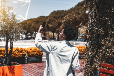 Kıvırcık Afrika saçlı, beyaz siperli, güneş gözlüklü, güneşli bir caddenin tepesinde kameralı telefonuyla fotoğraf çeken siyahi bir hippi kadının arkasına bakın.