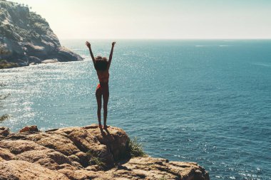 Kıvırcık saçlı, mayo giymiş, deniz mavisi bir okyanusun ve sonsuz ufkun önünde tropik bir tatil köyü sahilinin kenarında duran zarif, genç bir Afro-Amerikalı kadının silüeti.