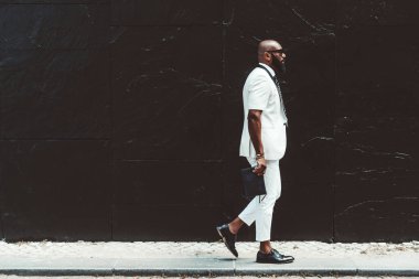 Beyaz kollu, kısa kollu, güneş gözlüklü ve deri ayakkabılı zarif sakallı bir Afrikalı adam elinde bir adamın debriyajı ile siyah bir duvarın önünden geçiyor.