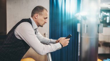 Ciddi bir iş adamı, ofis çalışma alanındaki balkon korkuluklarına yaslanırken akıllı telefonunu kullanarak çalışan bir yazışmaya cevap veriyor.