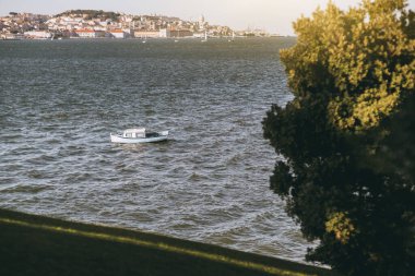Portekiz 'in Almada kentindeki Tejo nehrinin dalgalarında sallanan mavi çizgili perdeleri olan eski, küçük, rahat beyaz bir tekne. Arka planda, küçük, antika Lizbon evleriyle dolu bir nehir kıyısı.