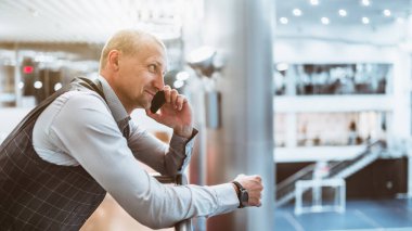 Olgun, neşeli girişimci bir adam gülümsüyor ve iş arkadaşıyla telefonda konuşurken modern ofis dinlenme alanının üzerindeki korkuluklara yaslanıyor.