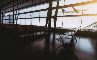 Boş, karanlık, çağdaş bir havaalanı bekleme salonu. Sıralar dolusu ahşap koltuklar ve arka planda irtifa kazanan bir yolcu uçağı. Geniş açılı, camdan bir ön cephede.