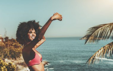 Arkasında güneşli bir okyanus olan tropikal bir tatil köyünün uçurumunda dururken cep telefonuyla neşeli bir video görüşmesi yapan göz kamaştırıcı bir Brezilyalı kadın.