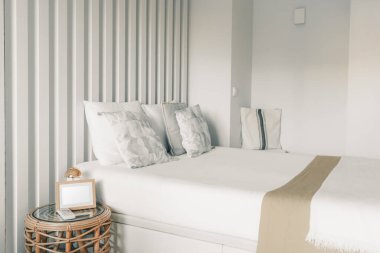 Üzerinde yastıklar ve yastıklar olan bir otel odasının yatak odasında beyaz bir yatak odası ve üzerinde boş bir çerçevenin beyaz şablonu olan küçük bir komodin.