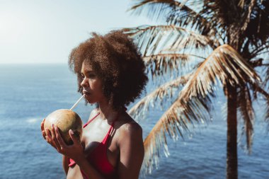 Okyanus kıyısındaki tropik bir tatil köyünün kıyısında dururken gerçek hindistan cevizi suyu içen kırmızı mayo giymiş genç ve susamış bir Afro-Amerikalı kadının portresi.