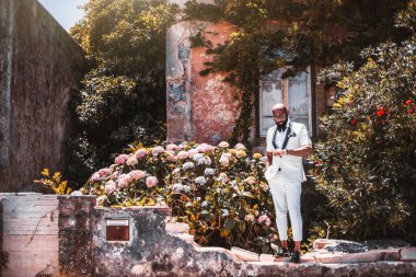 Süslü, kel, sakallı, beyaz takım elbiseli bir adam antika, terk edilmiş bir Avrupa evinin penceresi ve küçük bir bahçesi olan arka planında dikilirken saatine bakıyor.