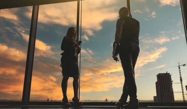 A conversation of two business partners on the top of an office skyscraper in the evening during a stunning sunset, near the window, selective focus on a businessman, his female colleague is defocused