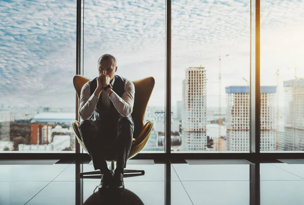 A portrait of a man entrepreneur in a state of worry and dismay sitting on an armchair in front of the window of a luxury office high-rise and thinking about how to solve recent business issues
