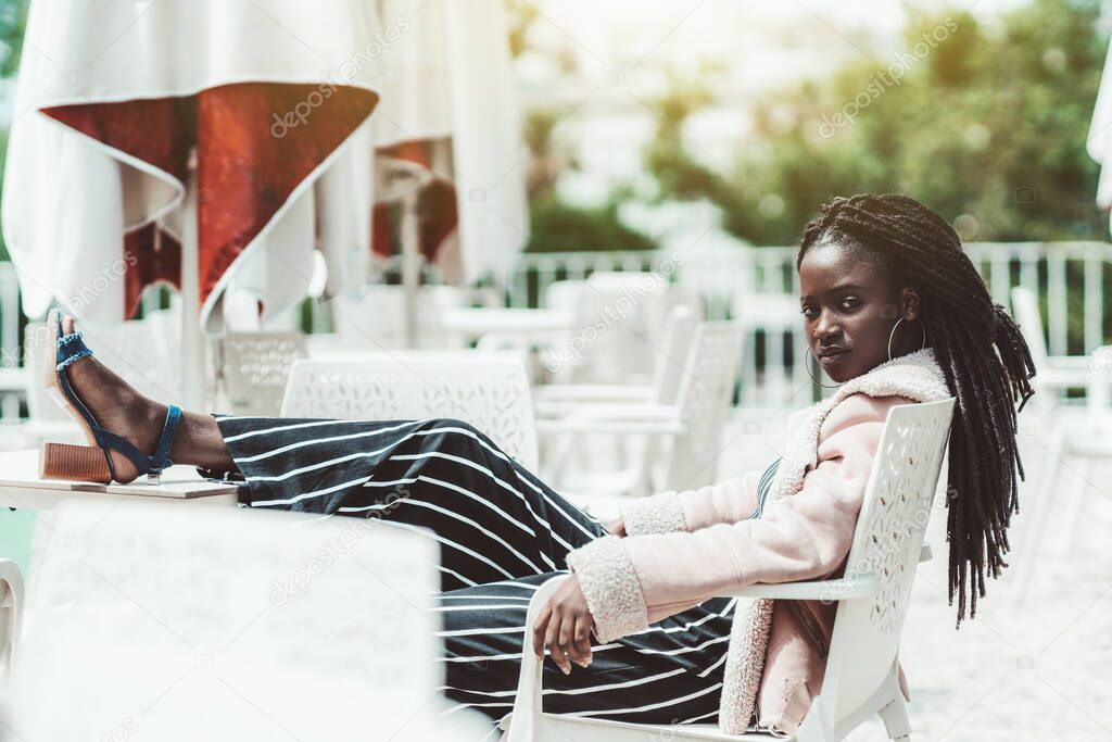 A portrait of a young ravishing black female with dreadlocks and earrings and in a demis-season coat, and striped trousers, lounging on a chair in an outdoor cafe with legs on the table, arrogant look