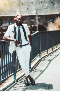 Zarif, kel, beyaz bir kostüm ve kırmızı kablosuz kulaklık takmış siyah bir adam sokakta müzik dinliyor; bir Afrikalı adam dışarıda metal bir çite yaslanıyor.