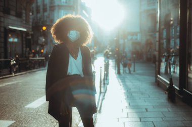 Kıvırcık saçlı, virüs koruması maskeli genç bir siyahi kadının silueti ve akşam güneşiyle aydınlanan yolun yakınındaki kaldırımda duran paltosu.