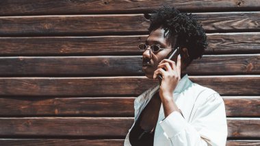 Ahşap bir yüzey önünde telefonla konuşan genç bir siyahi kadının profil portresi; güneş gözlüklü ve beyaz siperli büyüleyici Afrikalı kadın dışarıda akıllı telefondan sohbet ediyor.