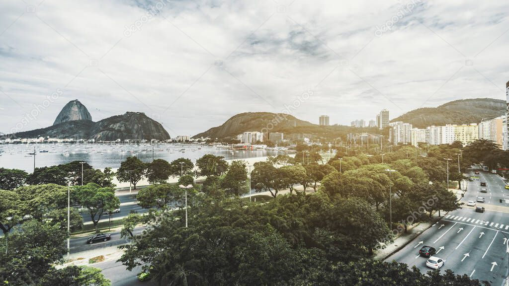Wide-angle cityscape of Botafogo district of Rio de Janeiro with multiple lane roads with cars, lines of trees, residential buildings, Sugar Loaf mountain, a bay with plenty of sailboats, overcast sky