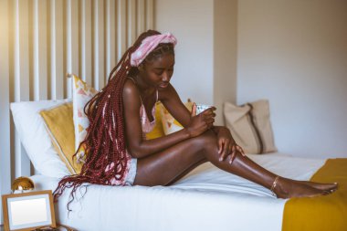 Göz kamaştırıcı siyah bir kadın uyandı ve elinde bir fincan çay ile yatağında serinliyor; uzun örgülü genç siyah bir kadın sabah yatakta oturuyor ve kahve içiyor