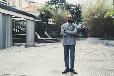 Zarif gözlüklü, şık, tüysüz, siyah, yakışıklı bir girişimci ve şık, özel yapım bir kostüm giymiş, metalik bir zeminde dikiliyor, yan yan bakıyor.