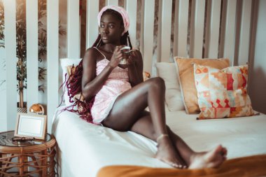 Düşünceli, genç, şehvetli, siyah ayaklı, gecelikli ve örgülü bir kadın, sıcak dairesinin yatak odasındaki yatakta soğuyor. Ellerinde lezzetli bir fincan çay tutarken yan bakıyor.