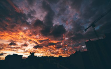 Arka planda eflatun ve turuncu renkte dramatik bir gün batımı olan çarpıcı geniş açılı şehir manzarası ve evlerin siluetleri ve şehrin ön planında bir inşaat vinci.