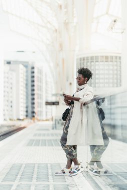 Güneş gözlüklü, beyaz siperli ve kamuflaj pantolonlu şık, şık, genç siyahi kadın, tren istasyonundaki bir tren istasyonunun cam çitine yaslanırken akıllı telefon kullanıyor;