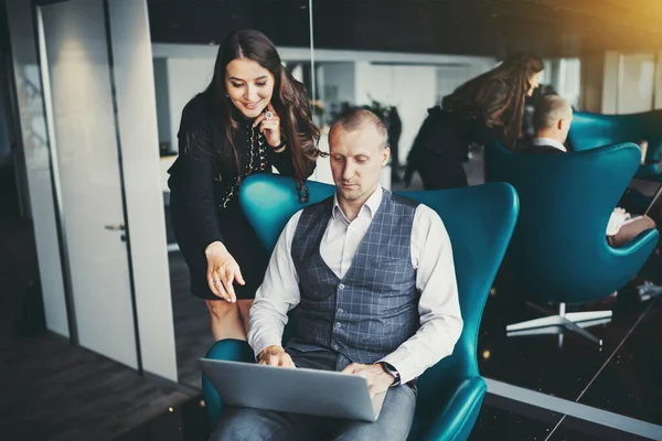 A business couple: a man entrepreneur on a teal armchair is sharing the screen of his laptop with his female colleague, a hispanic businesswoman who is standing near him and pointing on the screen