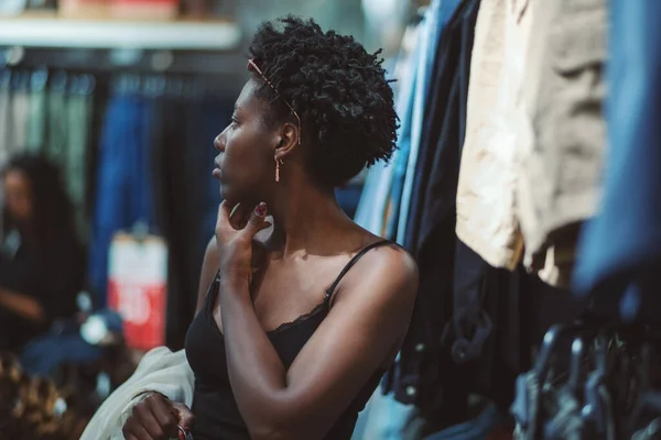 A beautiful young black woman with a hanger in hand is pensively looking around while standing in a clothing store with various textiles and clothes hanging around; shallow depth of field