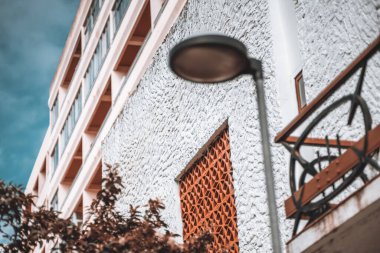 Kil pencere ızgaralı kaba desenli beyaz bir duvara seçici bir odak ile bir ev cephesi görüntüsü; bir sokak lambası ve yerinden çıkmış ön planda bir balkon parçası; sığ alan derinliği