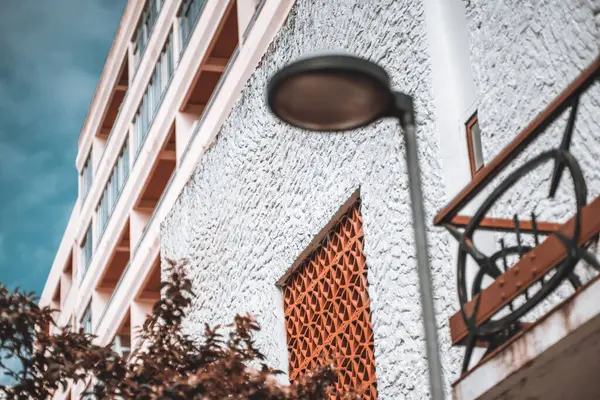 Kil pencere ızgaralı kaba desenli beyaz bir duvara seçici bir odak ile bir ev cephesi görüntüsü; bir sokak lambası ve yerinden çıkmış ön planda bir balkon parçası; sığ alan derinliği