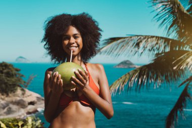 Kıvırcık saçlı, mayo giymiş, elinde pipetle elinde hindistan ceviziyle tropikal bir tatil köyünün önünde duran mutlu, genç, Afro-Amerikan bir kadının portresi.