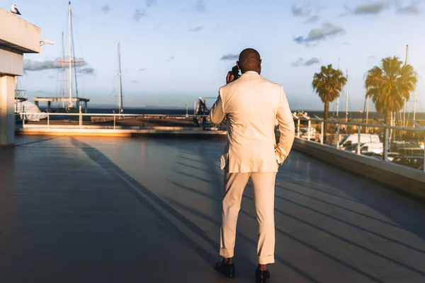 View from behind of a black guy in an elegant suit speaking on the phone while standing on a sunset coastal area; African bald man in a white costume taking on the phone outdoors, rear view