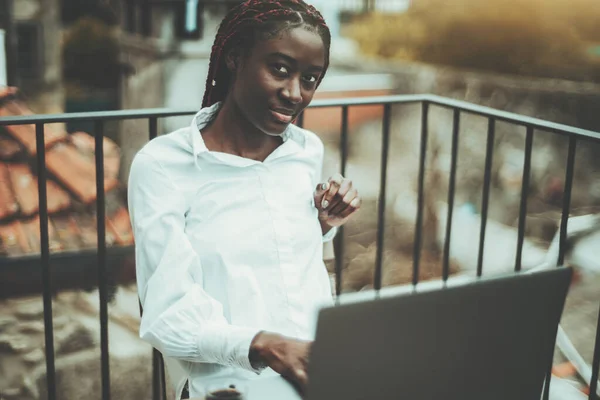 A portrait of a young beautiful black female freelancer with braided hair and in a white shirt, working with her freelance project using the laptop while sitting on the balcony of a street cafe