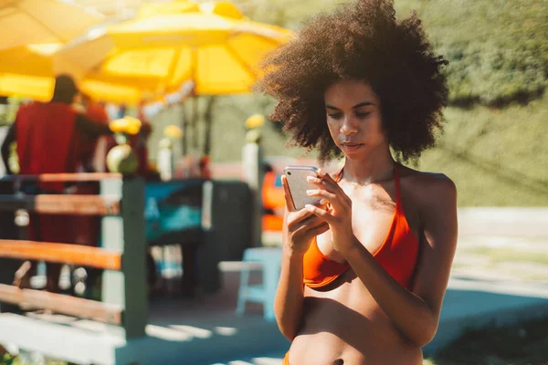 一个年轻迷人的非洲裔美国女人的画像 她长着蓬松的非洲裔头发 身穿泳衣 夏天站在海滩小摊边 通过智能手机向她的新男友传达了一个信息 — 图库照片