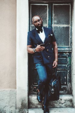 Süslü, kel, sakallı, papyon takmış, deri çantalı, tahta kapıdan çıktıktan sonra antik bir binanın verandasında duvara yaslanmış, kel ve olgun bir zenci.