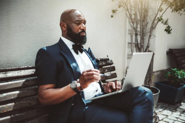 A fancy handsome bald bearded African senior with glasses in a hand is working on a netbook outdoors; portrait of a black man entrepreneur with a well-groomed beard using a laptop on a street bench