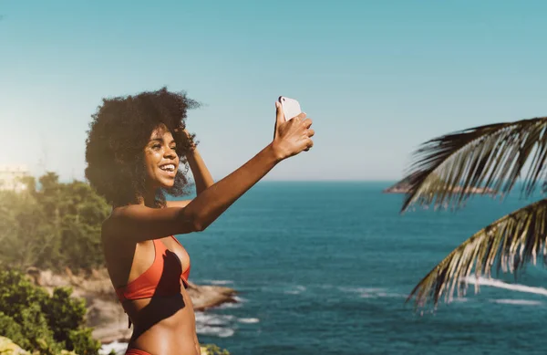 在热带旅游胜地 年轻的巴西女性穿着红色泳衣站在大海前 一边用智能手机播放视频 一边自拍 — 图库照片