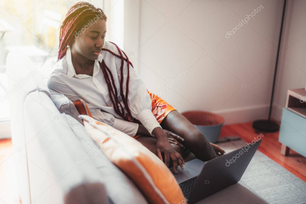Portrait of a ravishing young black female with braided red afro hair, sitting on the couch in a cozy interior of her living room and watching a series via her laptop; a copy space place on the right