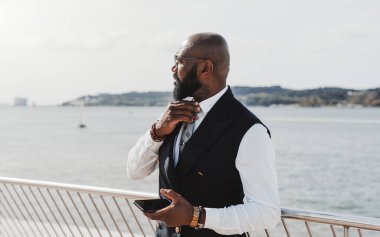 Gözlüklü ve takım elbiseli zarif sakallı siyah bir işadamı, arka planda bir nehir olan yüksek katlı bir iş yerinin çitinin yanında elinde cep telefonuyla kravatını düzeltiyor.