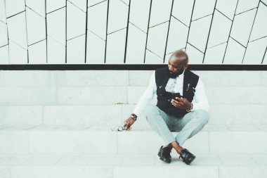 Süslü, kel, yelekli, şık bir Afrikalı adam elinde akıllı bir telefonla mermer bir basamakta oturuyor ve gözlüklerine uzanıyor. Solda bir fotokopi odası var.