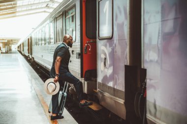 Zarif bir yaz elbisesi ve elinde bavul çantası ve şapkası olan süslü, kel, sakallı bir zenci tren istasyonundaki tren istasyonundaki yolculuğuna başlamak üzere trene biniyor.