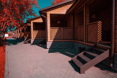 An infrared shot of a group of similar wooden houses of a youth hostel or a campsite with verandas and small stairs leading to stoops; a paving-stone track on the left next to trees with red leaves clipart