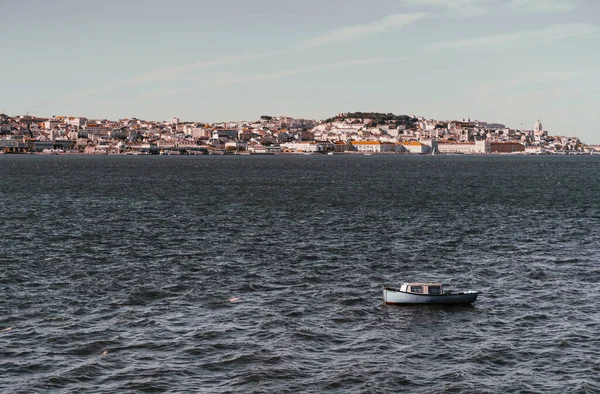 Kulübesinin içinde mavi kaplamalı ve çizgili perdeleri olan, Portekiz 'in Tejo nehrinin dalgalarında sallanan, arka planda antik Lizbon evleriyle dolu küçük bir nehir kıyısı olan küçük, rahat bir tekne.