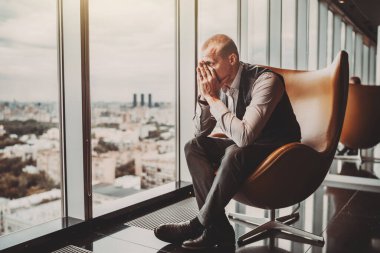 A stressed out adult caucasian man entrepreneur partly closing face with his hands while sitting on an office armchair next to the window of a modern business skyscraper and looking on the cityscape