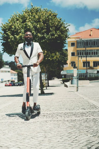 A vertical shot of a dapper adult bearded bald black senior in eyeglasses and a white fancy summer costume is holding in hands the handlebars of the e-scooter ready to ride over the paving-stone